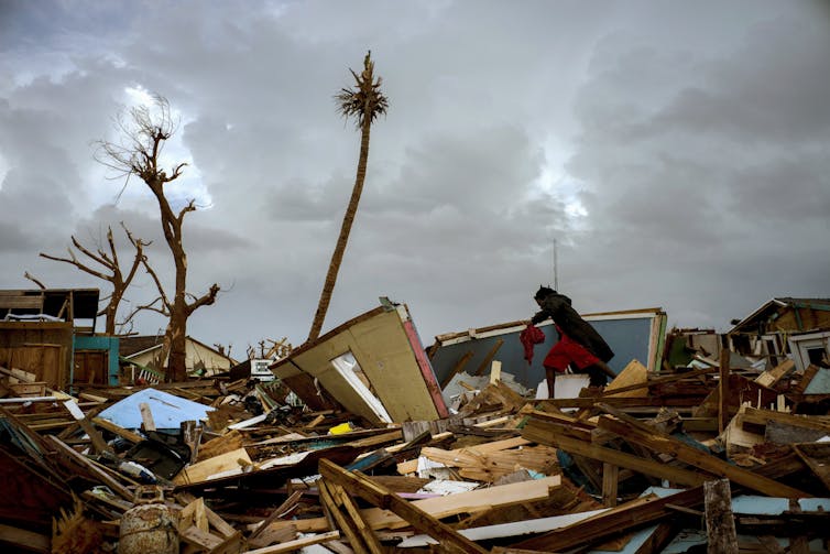 A person jumps over debris next to what remains of a home. Its roof is missing, and the walls are askew.