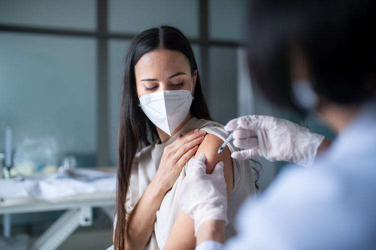 A young woman wearing a face mask receives a vaccination in her upper arm.