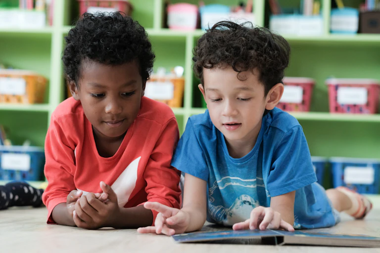 Two boys are seen lying on the ground looking at a book.