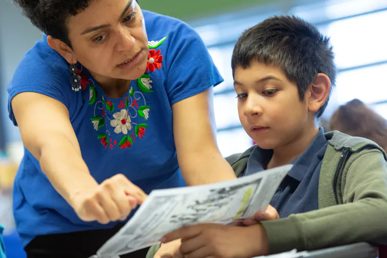 A teacher is seen beside a boy; they look at text.