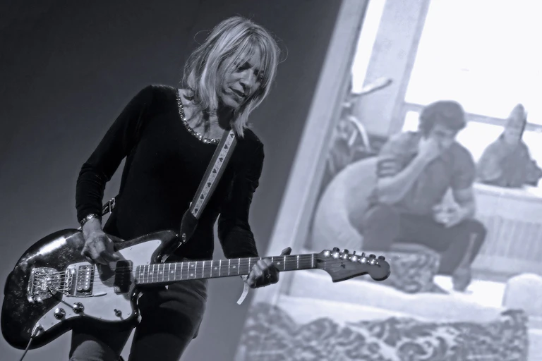 Woman playing a guitar, with a man behind her in hotel room, sitting on a chair