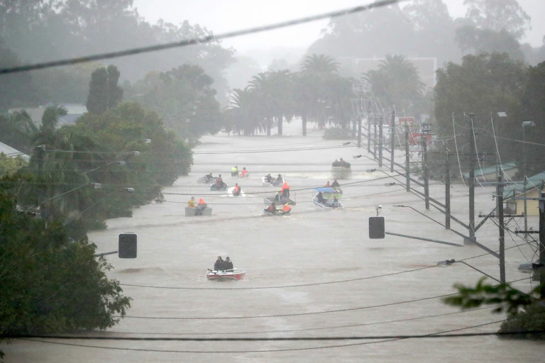 Photo showing people in boats floating on a heavily flooded street, with water almost reaching traffic lights and power lines.