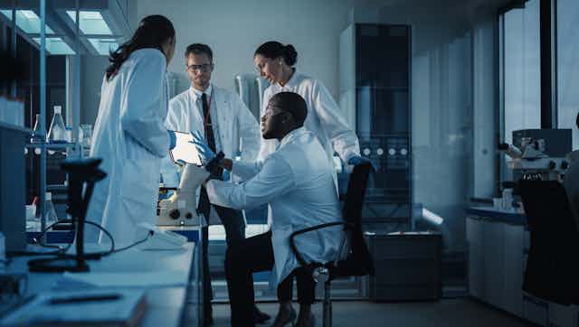 Researchers in a lab, wearing white coats, look over a screen and discuss something