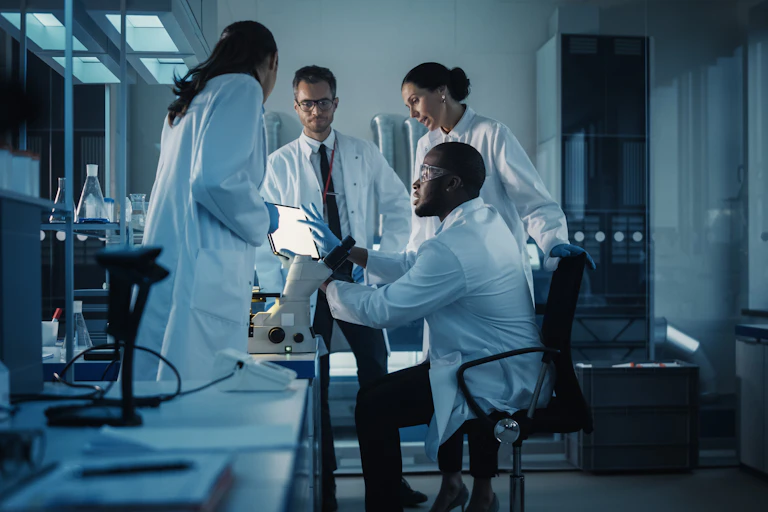 Researchers in a lab, wearing white coats, look over a screen and discuss something
