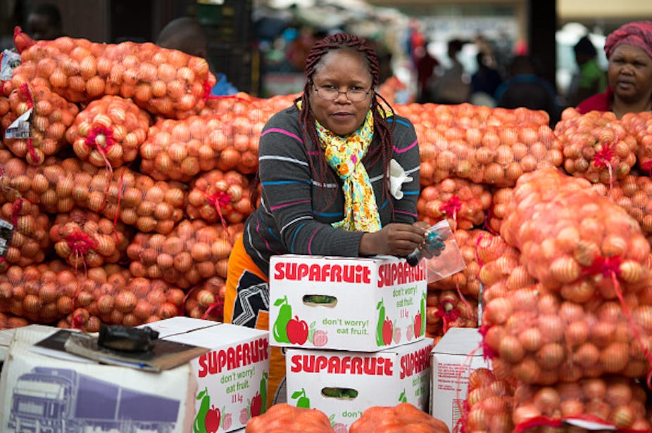 A woman is shown among bags of onions while leaning on boxes of fruit.