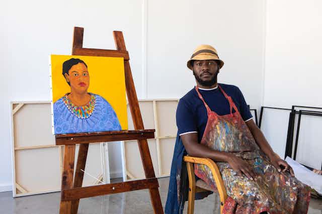 A man sits in a chair copying the pose and expression of a woman in a painting on an easel next to him.