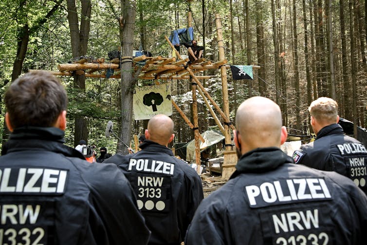 Police approach a wooden barricade in a forest.