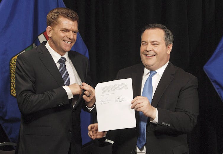 Two men in suits stand beside each other, one of them holding a document.