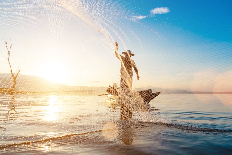 A person standing in a small boat throws a fishing net into the ocean.