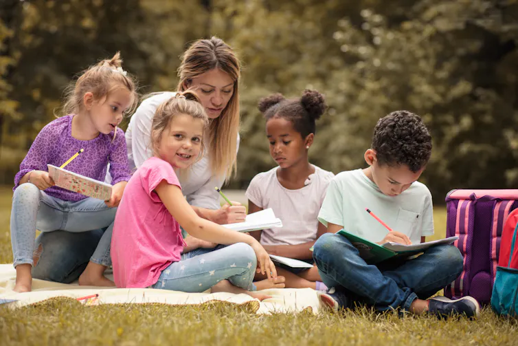 A teacher is seen with students sitting on the ground and they're holding papers and pencils.