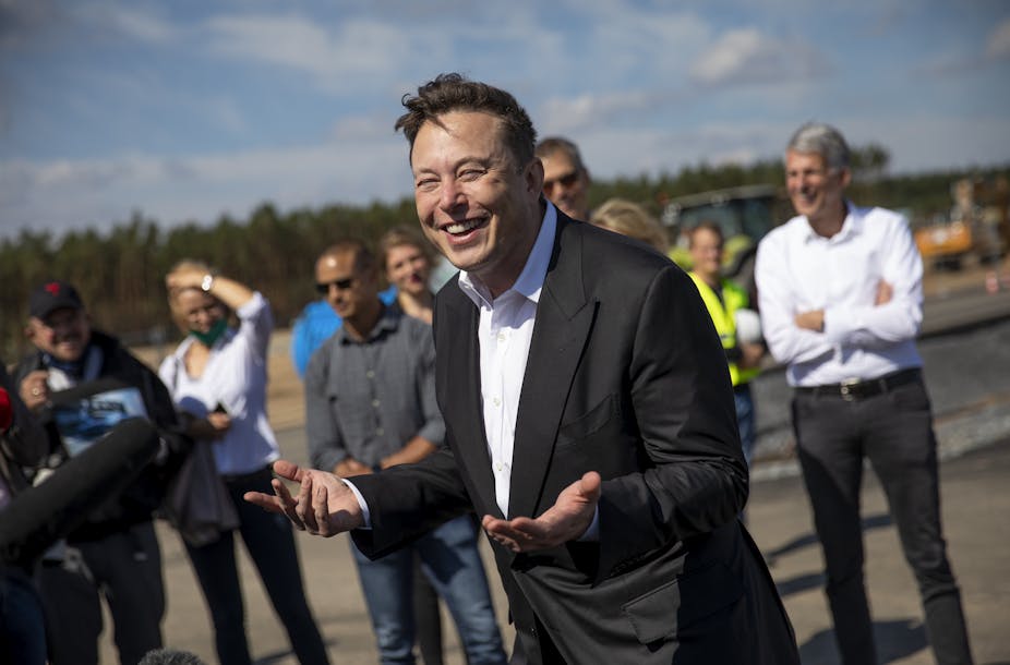 Elon Musk standing outside with people behind him holds out his hands, palms up, and has an amused look on his face.