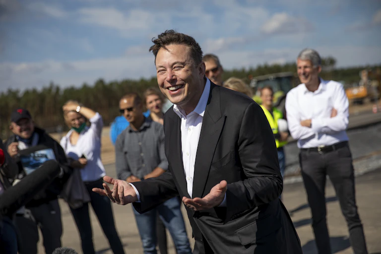 Elon Musk standing outside with people behind him holds out his hands, palms up, and has an amused look on his face.