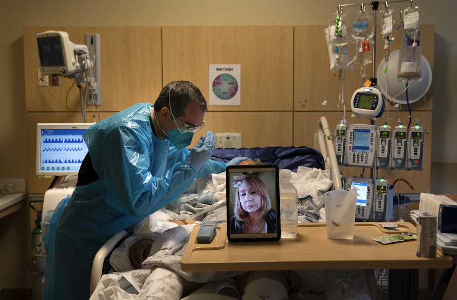 A chaplain in blue protective gear raising his hands to pray for a patient while the patient's daughter watches on video.