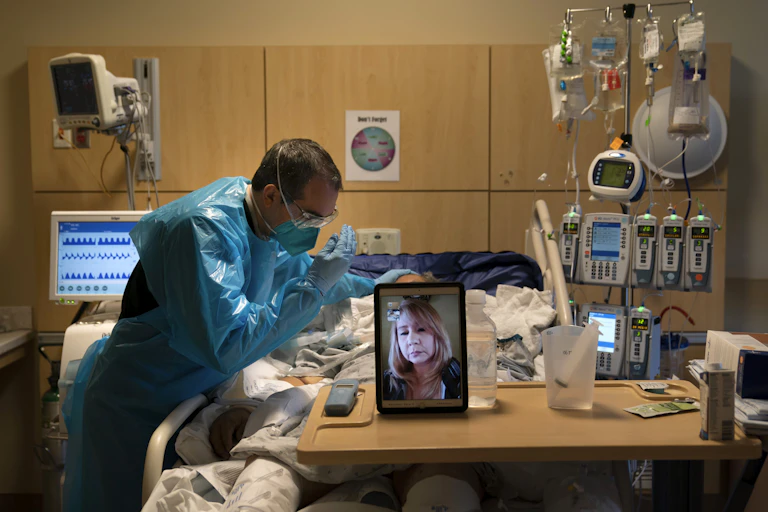 A chaplain in blue protective gear raising his hands to pray for a patient while the patient's daughter watches on video.