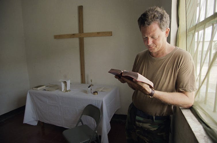 A man standing in front of a crucifix reading the Bible.