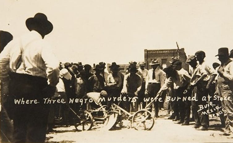 Dozens of men wearing hats have their heads down as they look at the site where three black men were burned at the stake.