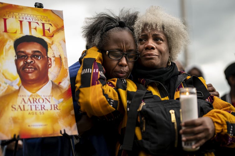 Dos mujeres negras se abrazan y se paran junto a un cartel de celebración de la vida en honor a Aaron Salter Jr., un hombre negro asesinado durante el tiroteo masivo en Buffalo.