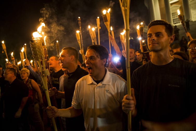 White men crowd together and appear to shout while carrying torches on a dark night,