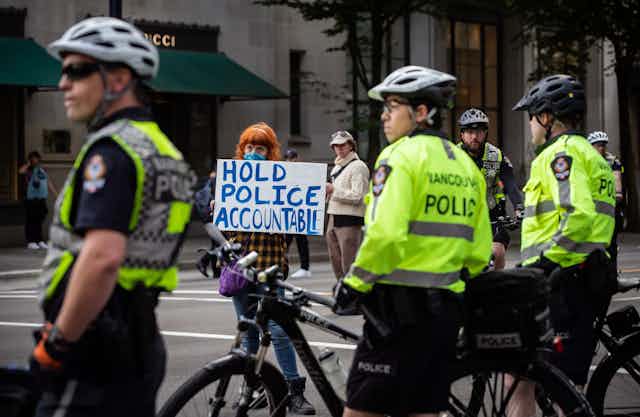 A woman holds a sign reading "Hold Police Accountable" near police officers watching