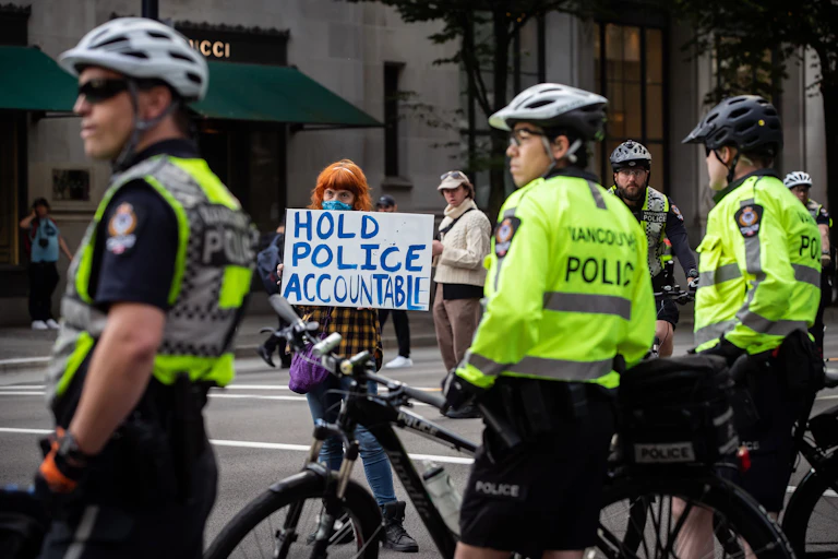 A woman holds a sign reading "Hold Police Accountable" near police officers watching