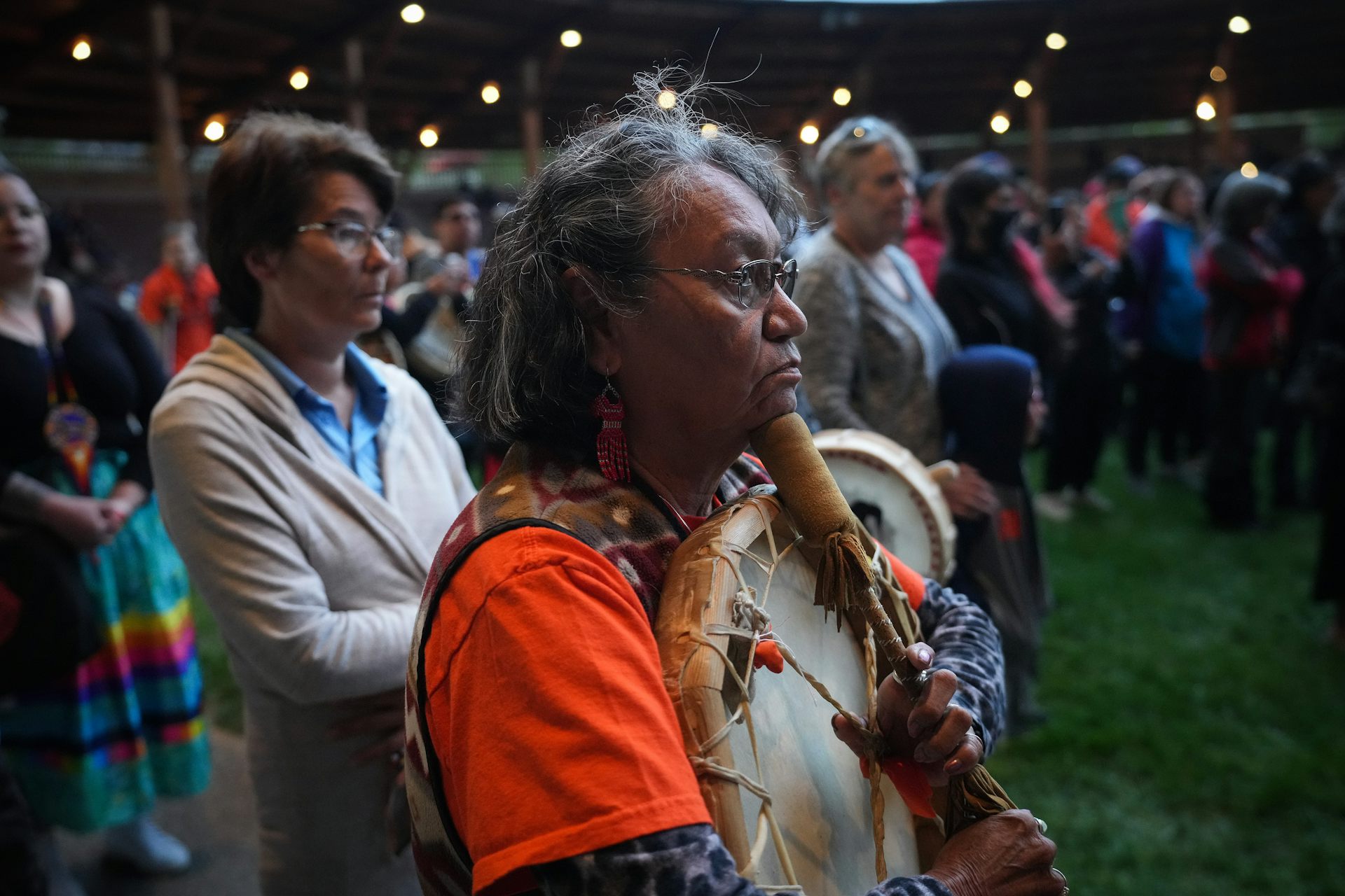 People stand looking solemn, a woman in an orange shirt clutches a hand drum.