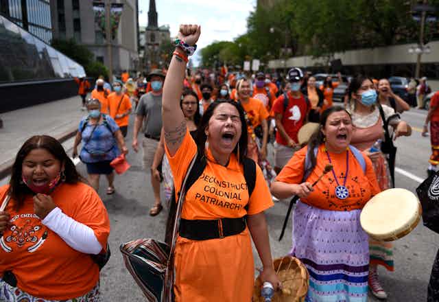A woman screams with her fist held in the air, she's wearing an orange shirt that reads 'deconstruct colonial patriarchy'