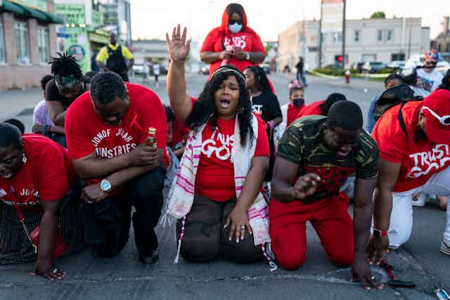 A small group of Black people kneel and put their heads down, except for the woman in the center who raises her hand up in prayer.