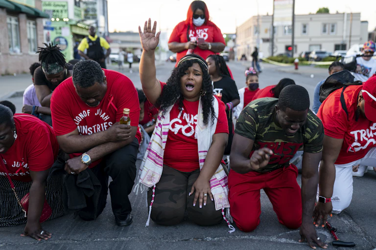 A small group of Black people kneel and put their heads down, except for the woman in the center who raises her hand up in prayer.