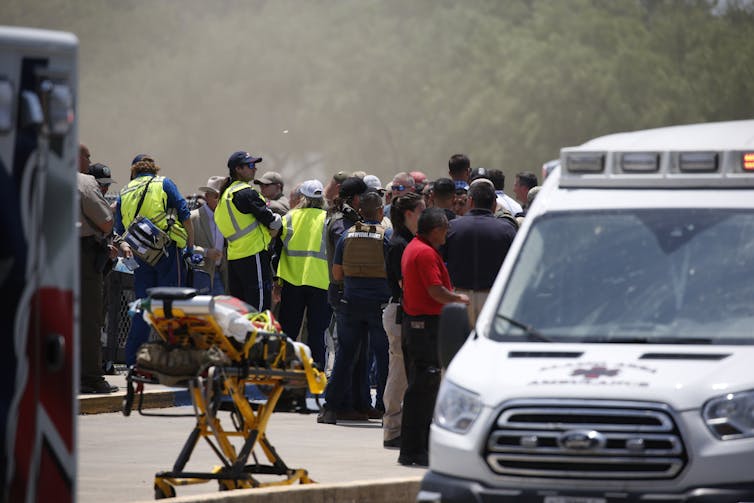 A crowd of people in uniforms and safety vests, standing near an ambulance and empty gurney.