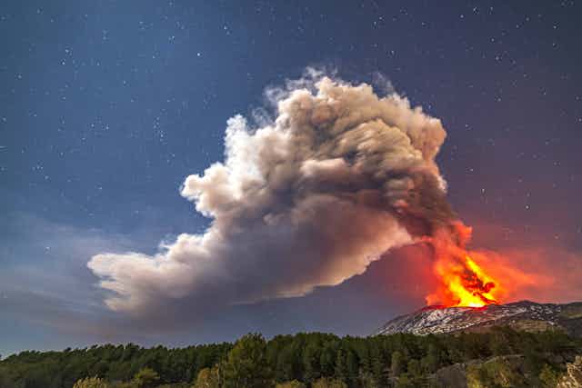 A nighttime photo showing a glowing volcano emitting a plume of smoke 