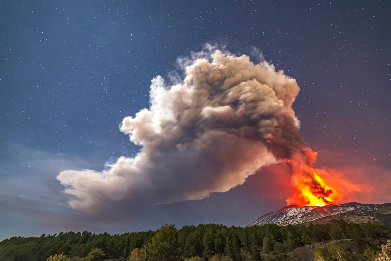 A nighttime photo showing a glowing volcano emitting a plume of smoke
