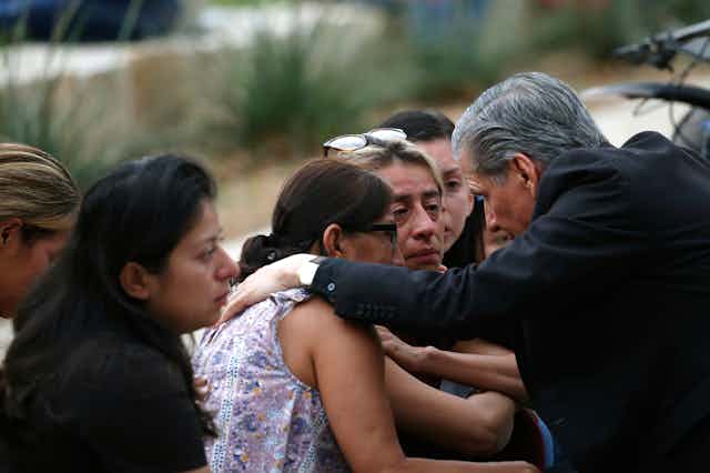 A gray-haired man in a black blazer comforts tearful women outside.