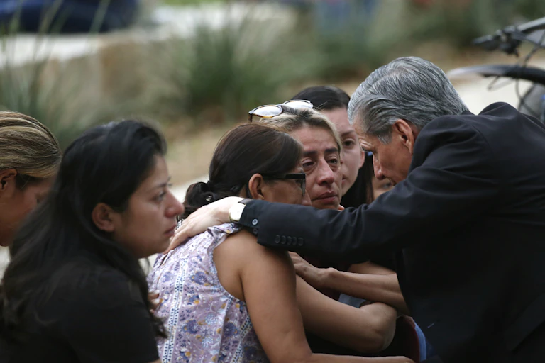 A gray-haired man in a black blazer comforts tearful women outside.