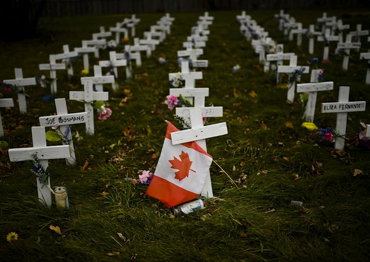 A row of white crosses on a green lawn. A small Canadian flag is attached to one of the crosses.