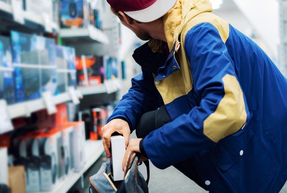 A man in an electronics shop squats and discreetly places an electronics box into his backpack, while looking away from the camera over his shoulder