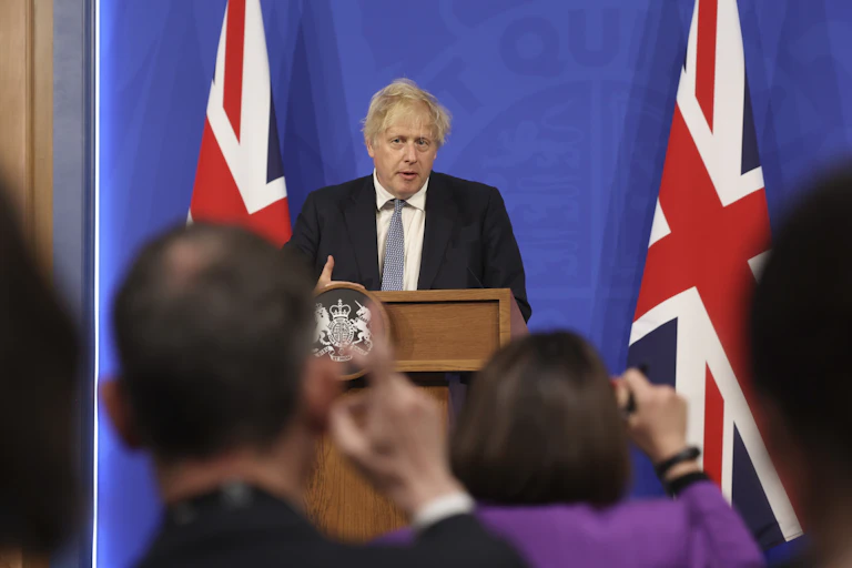Boris Johnson at a podium with members of the media raising their hands to ask him questions in the foreground.