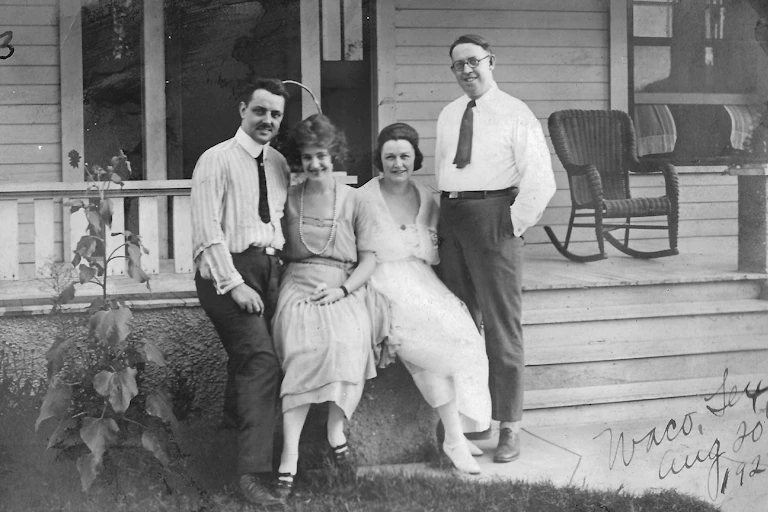 A photograph of a two smiling white couples standing next to each other near a front porch.