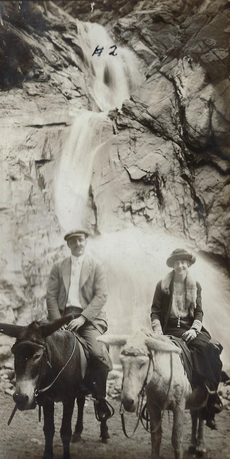 A  white man and woman are seen riding on donkeys near a mountain waterfall.