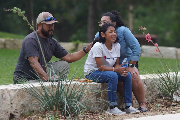 A girl in a white shirt cries, holding a water bottle, while she is comforted by a man and a woman.