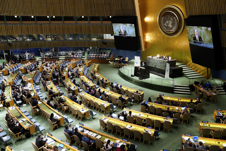 A man is seen on a screen addressing a large gathering in a cavernous meeting room.