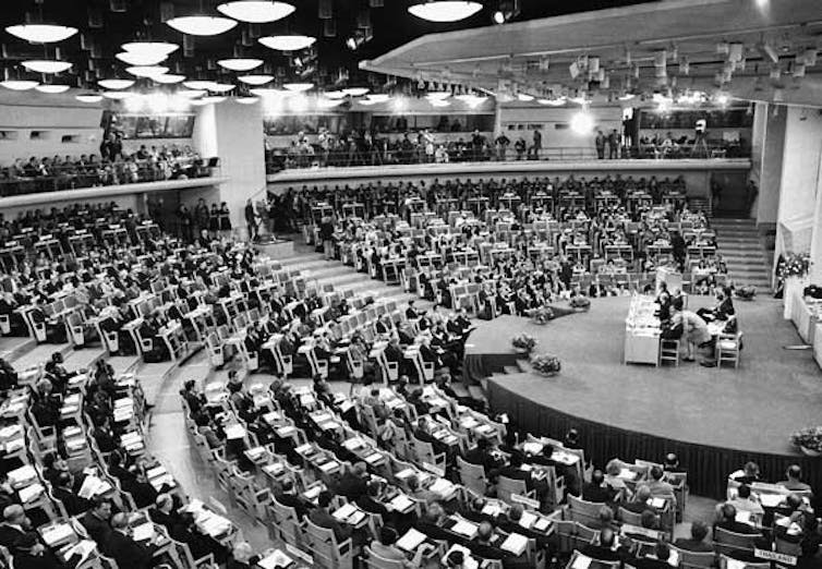 A conference hall filled with seated people and a person at the podium in the front.