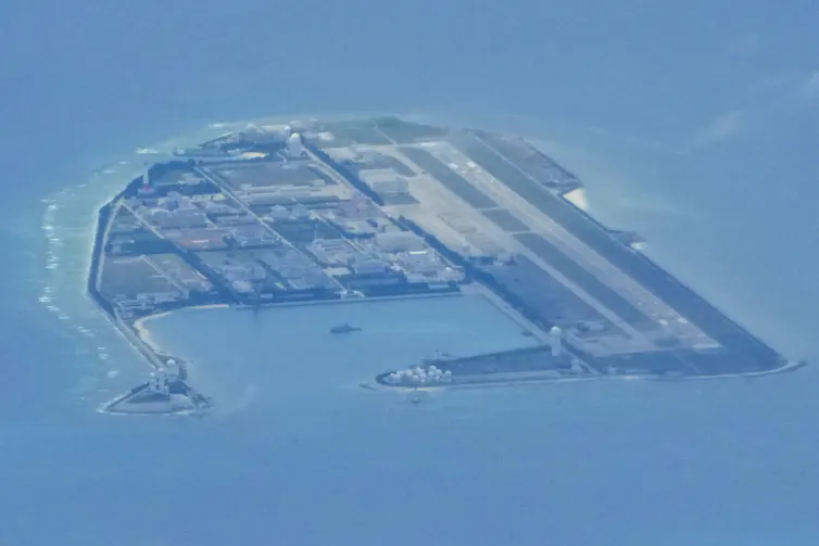 An island is seen from above with structures and buildings on it, surrounded by blue water.