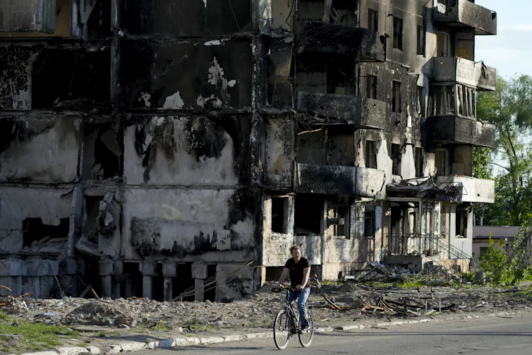 A man rides a bicycle in front of a building ruined by shelling.