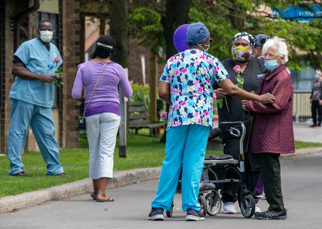 Long-term care workers in scrubs chat with an elderly resident outside a long-term care home.