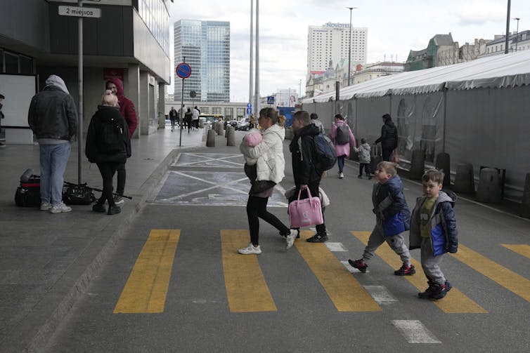 Uma mulher atravessa uma faixa de pedestres seguida por duas crianças do lado de fora de uma estação de trem.