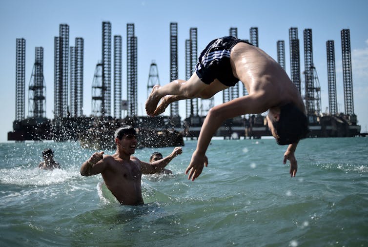 Teenagers play in the water of the Caspian sea - one young man is flipping another, with several Soviet oil rigs behind them.