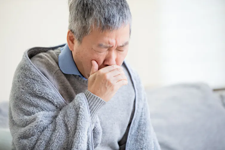 Older man draped in a blanket coughs into his hand.