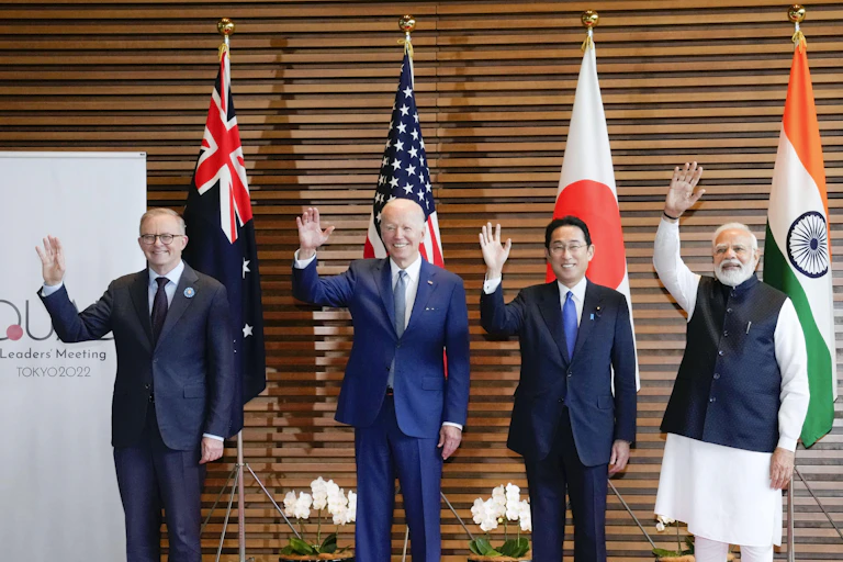 Four Quad leaders waving to the camera in front of their nation flags
