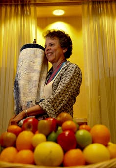 A woman holds a large scroll behind a tray of brightly colored fruit.