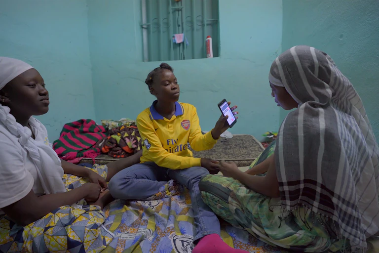 Three women sit cross-legged on the floor in a room with light blue walls. A young woman holds up a cell phone and shows the screen to a woman next to her.